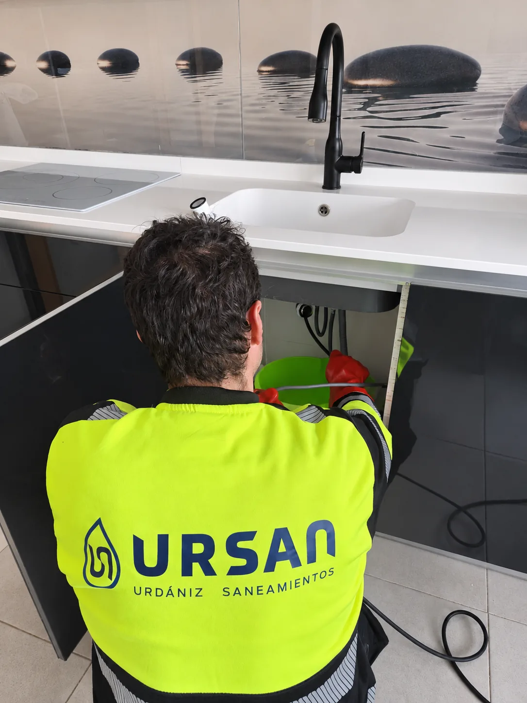 Profesional con ropa rotulada de URSAN trabajando en las tuberías bajo un fregadero de cocina.