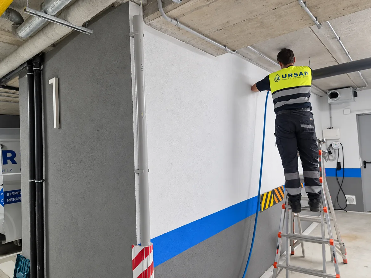 Profesional con ropa rotulada de URSAN sobre una escalera junto a las tuberías de un garaje.